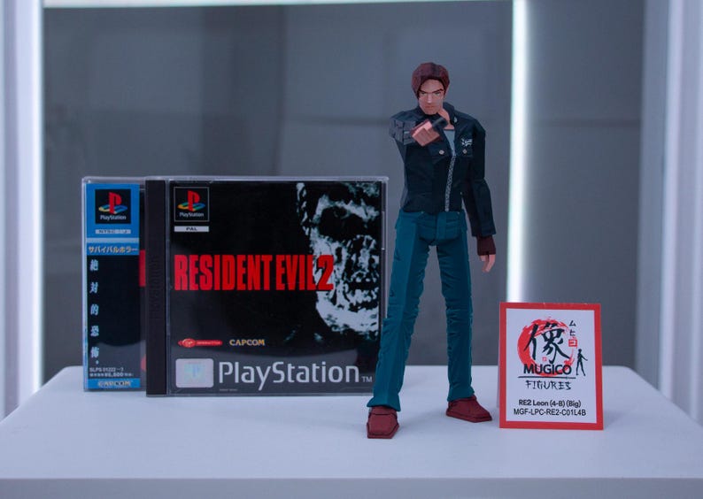 May include: A video game case for Resident Evil 2 for the Playstation console. The case is black with red text and a zombie on the cover. A red and white action figure of Leon S. Kennedy from the game is standing in front of the case. The action figure is holding a gun and has a serious expression on his face. A red and white card with Japanese text and the words "MUGICO FIGURES" is in the foreground.