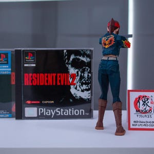 Puede incluir: Una figura de acci&oacute;n de una mujer con el pelo rojo, que lleva una chaqueta vaquera azul y pantalones vaqueros, sosteniendo una pistola. La figura est&aacute; de pie frente a una caja de juego de video para Resident Evil 2 para Playstation.
