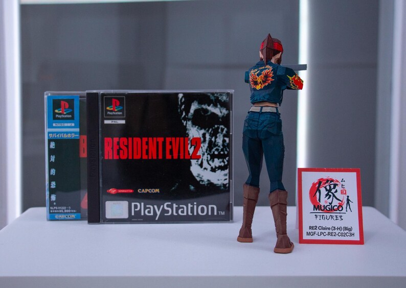 Puede incluir: Una figura de acci&oacute;n de una mujer con el pelo rojo, que lleva una chaqueta vaquera azul y pantalones vaqueros, sosteniendo una pistola. La figura est&aacute; de pie frente a una caja de juego de video para Resident Evil 2 para Playstation.