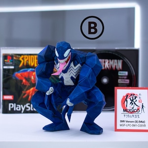 May include: A blue Venom figure in a crouching pose, with a white spider emblem on its chest and a pink tongue. The figure is in front of a Spider-Man PlayStation game case and a small red and white Mugico Figures card.