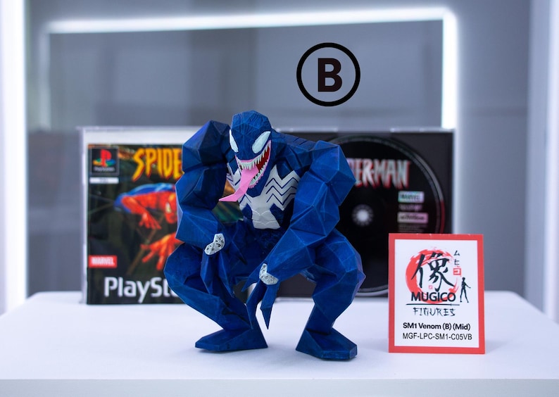 May include: A blue Venom figure in a crouching pose, with a white spider emblem on its chest and a pink tongue. The figure is in front of a Spider-Man PlayStation game case and a small red and white Mugico Figures card.