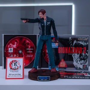 May include: A plastic action figure of a man in a black leather jacket and blue jeans holding a gun. The figure is standing on a base with the words "Resident Evil 2" printed on it. The figure is in front of two video game cases, one for "Resident Evil 2" and one for "Biohazard 2".