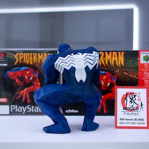 May include: A blue Venom figure with a white spider emblem on its back, posed in front of a Spider-Man PlayStation game box and a Nintendo 64 game box. A small card with Japanese text is also present.