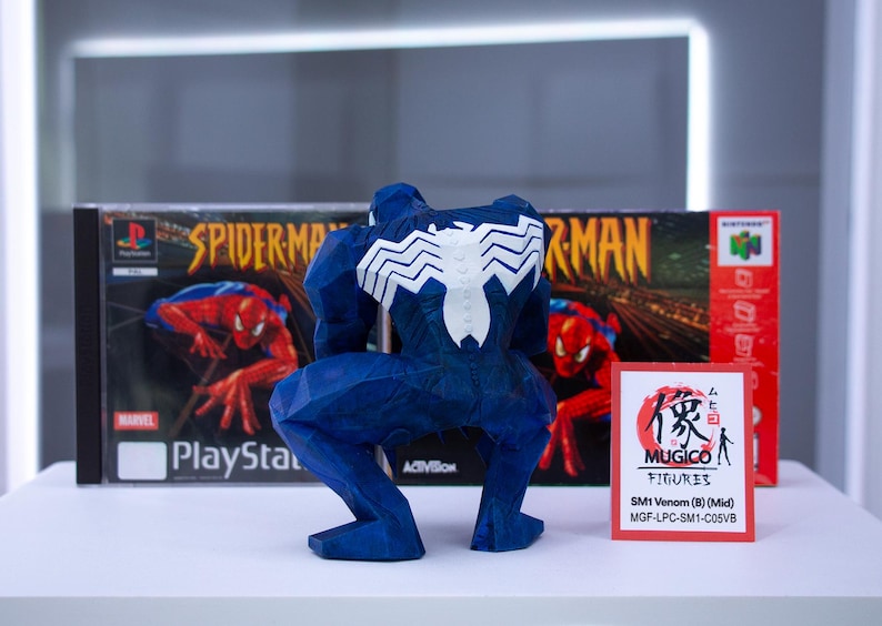 May include: A blue Venom figure with a white spider emblem on its back, posed in front of a Spider-Man PlayStation game box and a Nintendo 64 game box. A small card with Japanese text is also present.