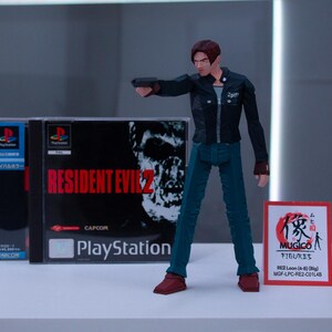 May include: A plastic action figure of Leon S. Kennedy from the Resident Evil 2 video game. The figure is wearing a black jacket, blue jeans, and brown boots. The figure is holding a gun in his right hand. The figure is standing in front of a video game case for Resident Evil 2. The case is blue and white and has the game title on it. The case is also labelled "PlayStation".