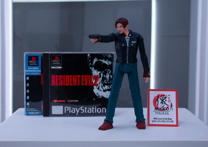 May include: A plastic action figure of Leon S. Kennedy from the Resident Evil 2 video game. The figure is wearing a black jacket, blue jeans, and brown boots. The figure is holding a gun in his right hand. The figure is standing in front of a video game case for Resident Evil 2. The case is blue and white and has the game title on it. The case is also labelled "PlayStation".