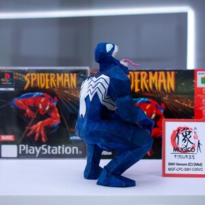 Puede incluir: Una figura azul de Venom con un emblema de ara&ntilde;a blanca en la espalda, agachada frente a una caja de juego de Spider-Man para PlayStation y una caja de juego roja de Nintendo 64. Un peque&ntilde;o letrero dice "MUGICO FIGURES".