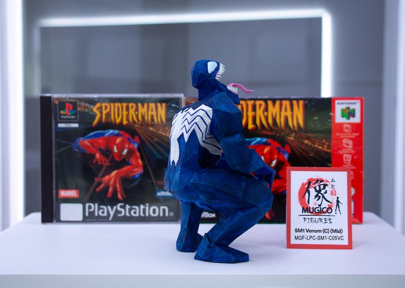 Puede incluir: Una figura azul de Venom con un emblema de ara&ntilde;a blanca en la espalda, agachada frente a una caja de juego de Spider-Man para PlayStation y una caja de juego roja de Nintendo 64. Un peque&ntilde;o letrero dice "MUGICO FIGURES".