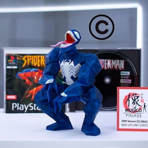 Puede incluir: Una figura azul de Venom con un emblema de ara&ntilde;a blanco, boca abierta y lengua larga, posando frente a una caja de juego de PlayStation de Spider-Man y una caja de CD. Una peque&ntilde;a tarjeta dice "MUGICO FIGURES".