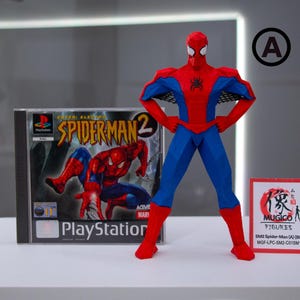 May include: A red and blue Spider-Man action figure stands next to a PlayStation 2 game case for Spider-Man 2. A small sign with Japanese text is also present. The figure is in a heroic pose.