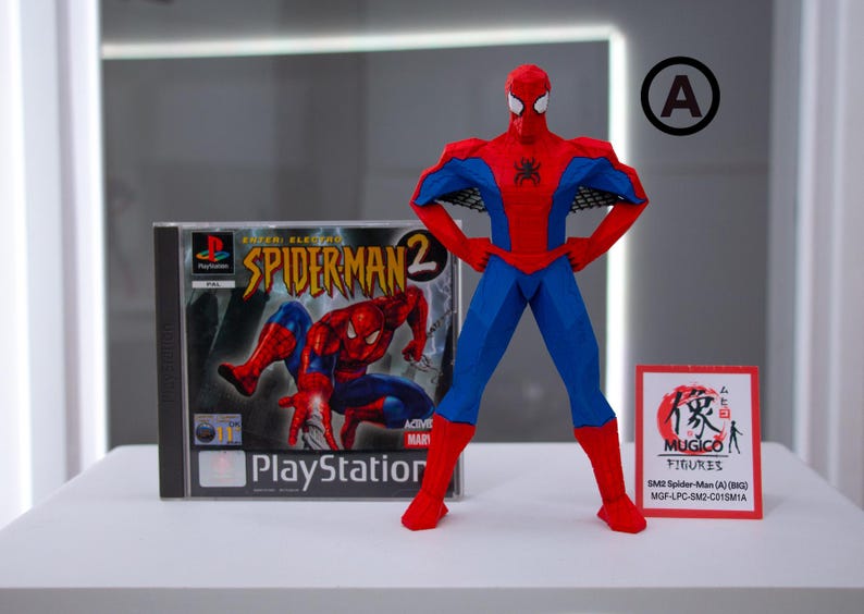 May include: A red and blue Spider-Man action figure stands next to a PlayStation 2 game case for Spider-Man 2. A small sign with Japanese text is also present. The figure is in a heroic pose.