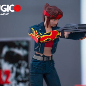 Puede incluir: Una figura de acci&oacute;n de pelo rojo que lleva una chaqueta vaquera azul con llamas amarillas y rojas en las mangas, un top rojo y unos vaqueros azules. La figura sostiene una pistola negra con la palabra "MUGICO" impresa en ella. La figura est&aacute; de pie sobre una superficie gris.