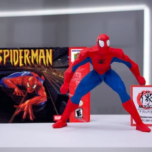 May include: A red and blue Spiderman action figure in a polygonal style, standing on a white surface. The figure is holding a small card with Japanese text and the words "MUGICO FIGURES" and "Spiderman (A) (Big) PC-SM1-C015PB01". The figure is in front of a video game box for the game "Spiderman" for the Nintendo 64.