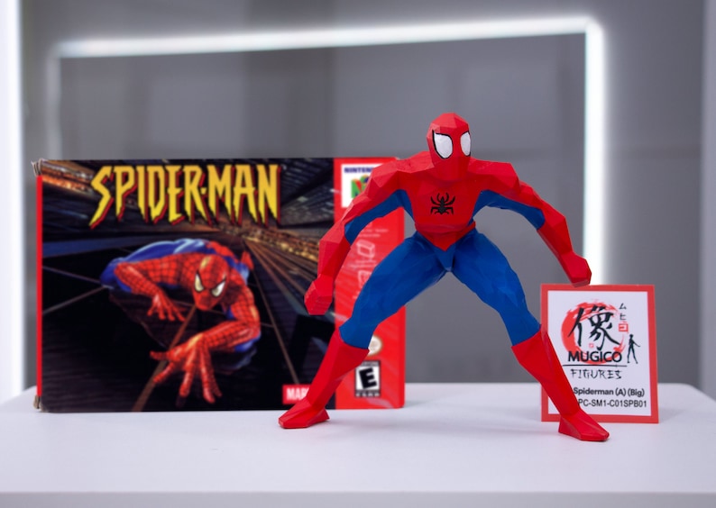 May include: A red and blue Spiderman action figure in a polygonal style, standing on a white surface. The figure is holding a small card with Japanese text and the words "MUGICO FIGURES" and "Spiderman (A) (Big) PC-SM1-C015PB01". The figure is in front of a video game box for the game "Spiderman" for the Nintendo 64.