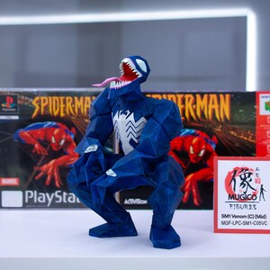 Puede incluir: Una figura azul de Venom con un emblema de ara&ntilde;a blanca, boca abierta y lengua larga, posando frente a una caja de juego de Spider-Man PlayStation y una caja de juego de Nintendo 64. Una peque&ntilde;a tarjeta roja dice "MUGICO FIGURES".