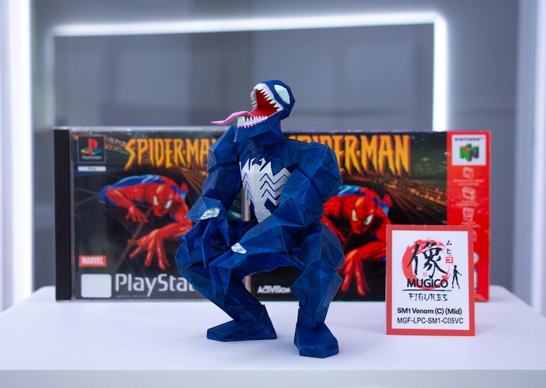 Puede incluir: Una figura azul de Venom con un emblema de ara&ntilde;a blanca, boca abierta y lengua larga, posando frente a una caja de juego de Spider-Man PlayStation y una caja de juego de Nintendo 64. Una peque&ntilde;a tarjeta roja dice "MUGICO FIGURES".