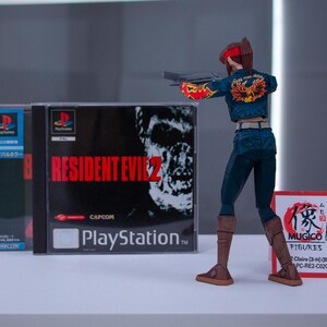 Puede incluir: Una figura de acci&oacute;n de una mujer pelirroja con un atuendo azul y rojo que sostiene una pistola. La figura est&aacute; de pie frente a una caja de juego de PlayStation para Resident Evil 2. La caja del juego tiene una cara de zombi en el frente. La figura lleva botas marrones.