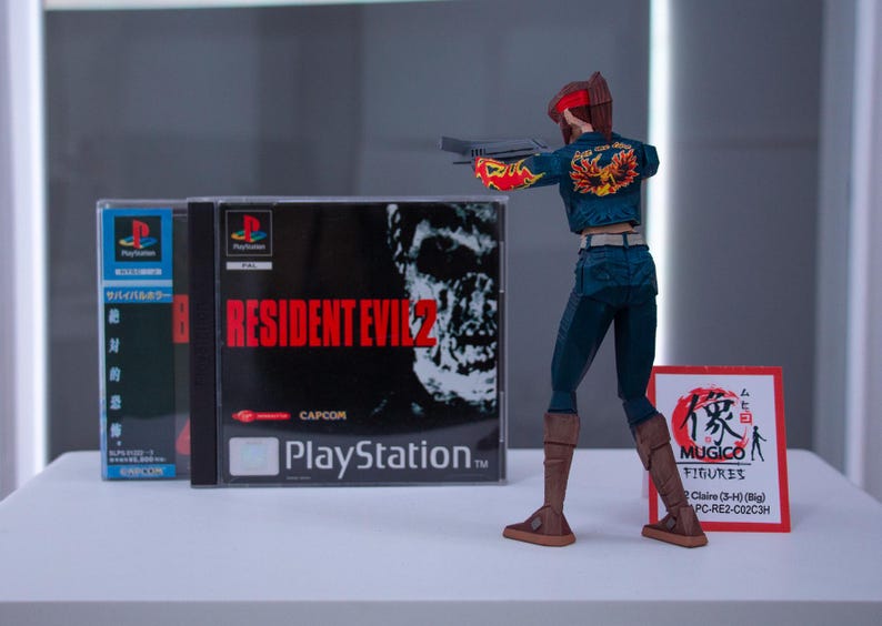 Puede incluir: Una figura de acci&oacute;n de una mujer pelirroja con un atuendo azul y rojo que sostiene una pistola. La figura est&aacute; de pie frente a una caja de juego de PlayStation para Resident Evil 2. La caja del juego tiene una cara de zombi en el frente. La figura lleva botas marrones.