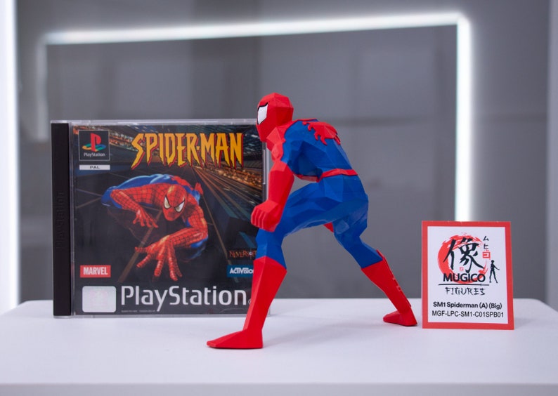 May include: A red and blue Spider-Man action figure in a crouching pose. The figure is made of a polygon style material. The figure is standing next to a PlayStation game case for Spider-Man. The game case has a red and blue Spider-Man on the cover. A small red card with Japanese text and the words "MUGICO FIGURES" is in the foreground. The card also has the text "SM1 Spiderman (A) (Big) MGF-LPC-SM1-C01SPB01".