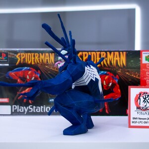May include: A blue Venom figure with white accents, posed in front of a PlayStation Spider-Man game box. The figure has a long tongue and spiky protrusions. A small sign reads "MUGICO FIGURES".