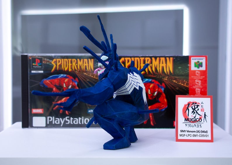 May include: A blue Venom figure with white accents, posed in front of a PlayStation Spider-Man game box. The figure has a long tongue and spiky protrusions. A small sign reads "MUGICO FIGURES".