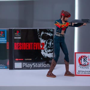 Puede incluir: Una figura de acci&oacute;n de una mujer pelirroja sosteniendo un arma, de pie frente a una caja de juego de Resident Evil 2 para Playstation. La caja tiene una cara de zombi en la parte delantera. La figura lleva una chaqueta roja y negra, pantalones vaqueros azules y botas marrones. La figura est&aacute; etiquetada como "MUGICO FIGURES RE2 Claire (3-4) (Big) MGF-LPC-RE2-C02C3H".