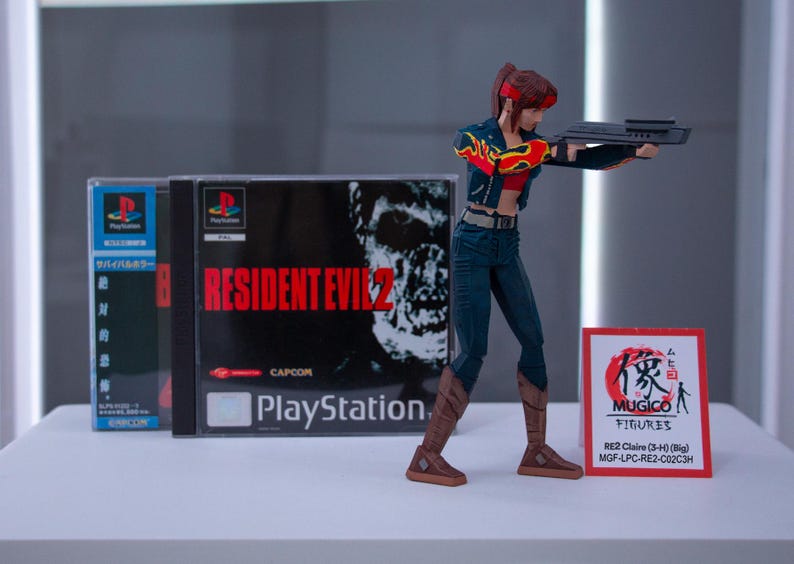 Puede incluir: Una figura de acci&oacute;n de una mujer pelirroja sosteniendo un arma, de pie frente a una caja de juego de Resident Evil 2 para Playstation. La caja tiene una cara de zombi en la parte delantera. La figura lleva una chaqueta roja y negra, pantalones vaqueros azules y botas marrones. La figura est&aacute; etiquetada como "MUGICO FIGURES RE2 Claire (3-4) (Big) MGF-LPC-RE2-C02C3H".