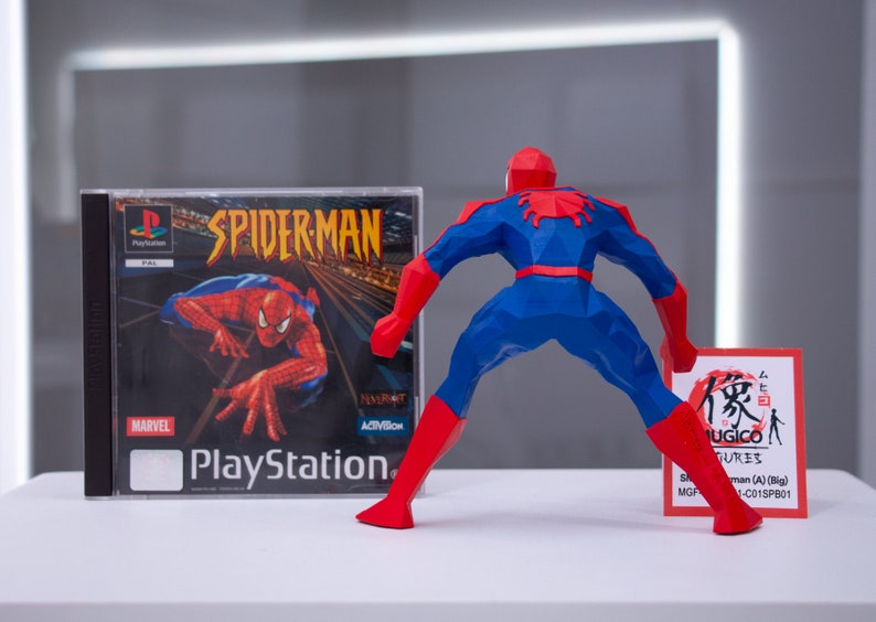 May include: A red and blue 3D printed figurine of Spider-Man standing with his back to the camera. The figurine is holding a small card with Japanese text that reads "Mugico Figures" and "Spider-Man (A) (Big)". The figurine is next to a PlayStation game case for Spider-Man.