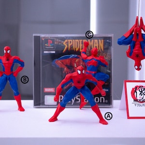 May include: A collection of Spider-Man figurines in red and blue, with a PlayStation game case in the background. One figurine is suspended upside down. A sign with Japanese text and the words "Mugico Figures" is also present.