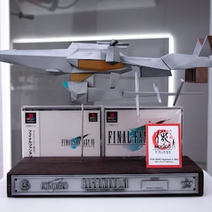 Pode incluir: Um modelo do dirigível Highwind de Final Fantasy VII, feito de papelão cinza e marrom, com duas caixas de jogos de PlayStation Final Fantasy VII e um adesivo MUGICO Figures. O modelo está em uma base de madeira com uma placa prateada que diz "FINAL FANTASY VII HIGHWIND 001".