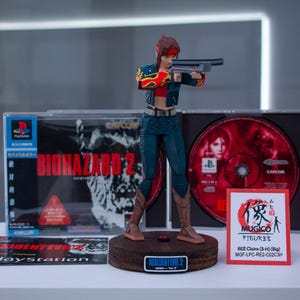 Puede incluir: Una figura de acci&oacute;n de una mujer sosteniendo un arma, vestida con ropa roja y negra. La figura est&aacute; de pie sobre una base de madera con las palabras "Resident Evil 2" impresas en ella. La figura est&aacute; rodeada de cajas de juegos de video para Resident Evil 2.