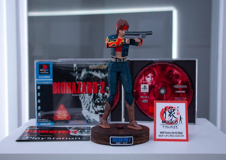 Puede incluir: Una figura de acci&oacute;n de una mujer sosteniendo un arma, vestida con ropa roja y negra. La figura est&aacute; de pie sobre una base de madera con las palabras "Resident Evil 2" impresas en ella. La figura est&aacute; rodeada de cajas de juegos de video para Resident Evil 2.