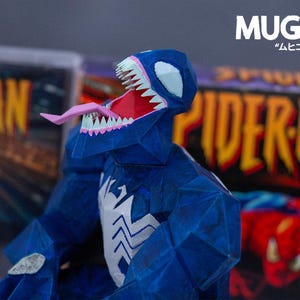 Puede incluir: Una figura de Venom azul de papel con la boca abierta, mostrando una lengua rosa y dientes blancos. La figura tiene un emblema de ara&ntilde;a blanco en el pecho. El fondo incluye portadas de c&oacute;mics con "Spider-Man" y "MUGIC".