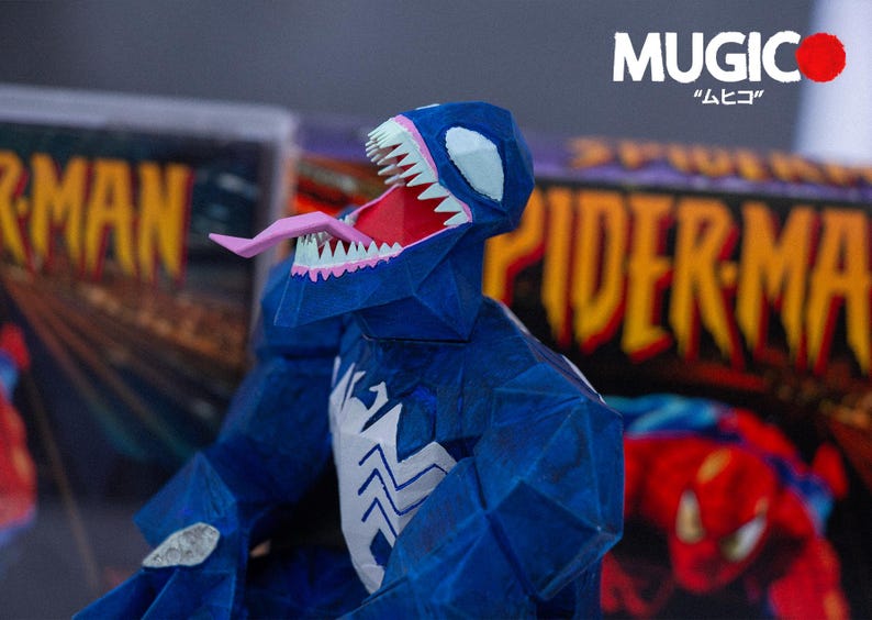Puede incluir: Una figura de Venom azul de papel con la boca abierta, mostrando una lengua rosa y dientes blancos. La figura tiene un emblema de ara&ntilde;a blanco en el pecho. El fondo incluye portadas de c&oacute;mics con "Spider-Man" y "MUGIC".
