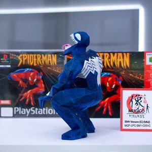 Puede incluir: Una figura azul de Venom hecha de papel, con un emblema de ara&ntilde;a blanco en la espalda, posando frente a una caja de juego de Spider-Man para PlayStation y una caja roja. Un peque&ntilde;o letrero rojo dice "MUGICO FIGURES".