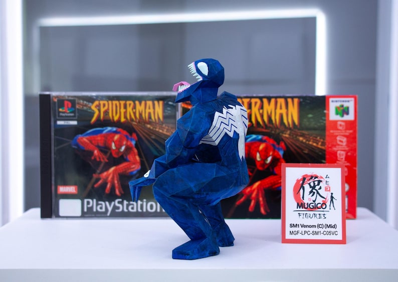 Puede incluir: Una figura azul de Venom hecha de papel, con un emblema de ara&ntilde;a blanco en la espalda, posando frente a una caja de juego de Spider-Man para PlayStation y una caja roja. Un peque&ntilde;o letrero rojo dice "MUGICO FIGURES".