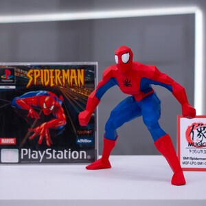 May include: A red and blue 3D printed action figure of Spiderman in a crouching pose. The figure is holding a small red and white card with Japanese text and the words "MUGICO FIGURES" printed on it. The card also includes the text "SM1 Spiderman (A) (Big) MGF-LPC-SM1-C015PB01". A Playstation game case for Spiderman is visible in the background.