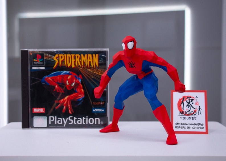 May include: A red and blue 3D printed action figure of Spiderman in a crouching pose. The figure is holding a small red and white card with Japanese text and the words "MUGICO FIGURES" printed on it. The card also includes the text "SM1 Spiderman (A) (Big) MGF-LPC-SM1-C015PB01". A Playstation game case for Spiderman is visible in the background.