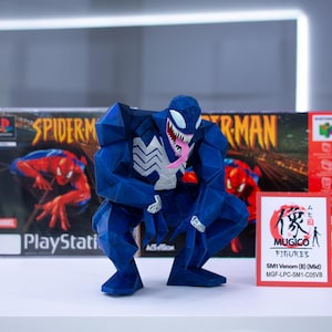 May include: A blue Venom figure with white detailing and a pink tongue, positioned in front of a Spider-Man PlayStation game box and a Nintendo 64 game box. A small card with Japanese text and "MUGICO FIGURES" is also visible.