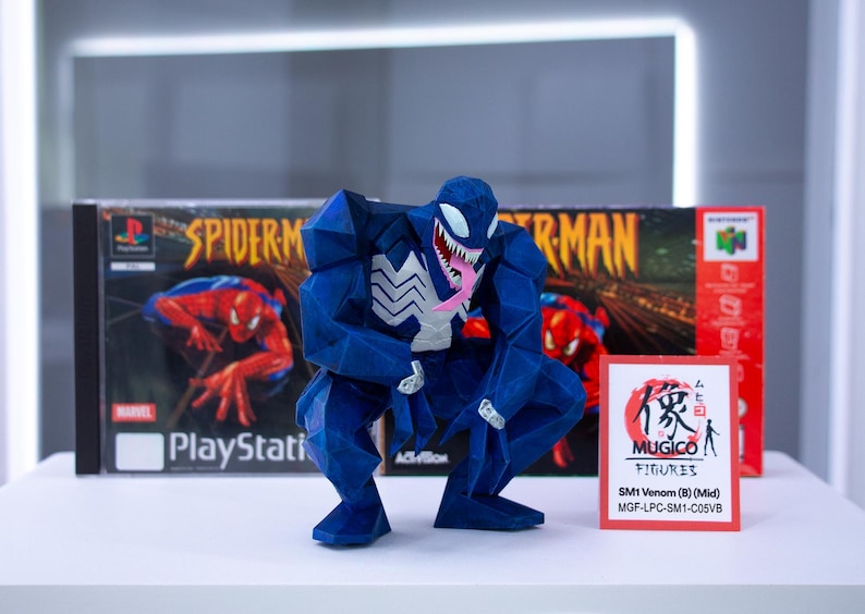May include: A blue Venom figure with white detailing and a pink tongue, positioned in front of a Spider-Man PlayStation game box and a Nintendo 64 game box. A small card with Japanese text and "MUGICO FIGURES" is also visible.