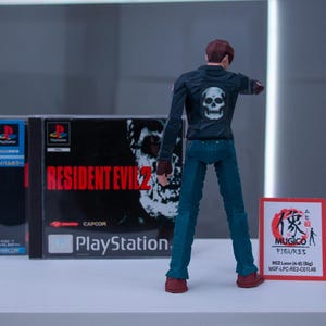 May include: A black and red video game case for Resident Evil 2 for the Playstation. A black action figure of a man wearing a black jacket with a skull on the back and blue jeans is standing in front of the case. A red and white card with Japanese text and the words "MUGICO FIGURES" is on the right side of the image.