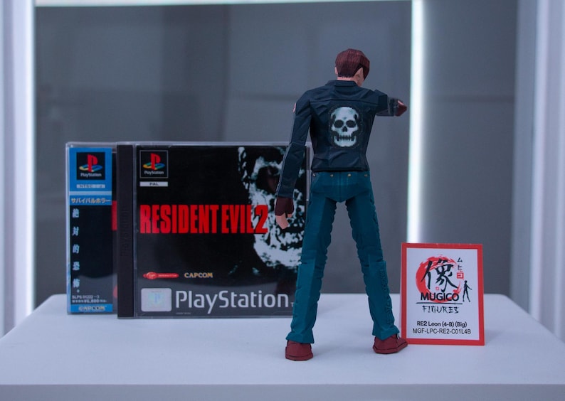 May include: A black and red video game case for Resident Evil 2 for the Playstation. A black action figure of a man wearing a black jacket with a skull on the back and blue jeans is standing in front of the case. A red and white card with Japanese text and the words "MUGICO FIGURES" is on the right side of the image.