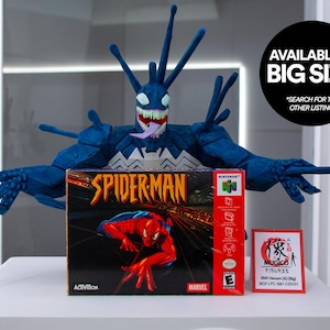 May include: A blue Venom figure with a white and black design, posed above a Spider-Man Nintendo 64 game box. The box is red and black with a Spider-Man graphic. A small red and white Mugico figure box is also present. Text on the image says "AVAILABLE IN BIG SIZE".