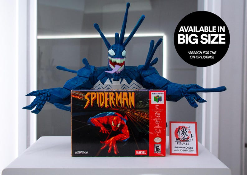 May include: A blue Venom figure with a white and black design, posed above a Spider-Man Nintendo 64 game box. The box is red and black with a Spider-Man graphic. A small red and white Mugico figure box is also present. Text on the image says "AVAILABLE IN BIG SIZE".