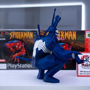 May include: A blue Venom figure with white accents and a long tongue, posed in front of a PlayStation Spider-Man game case. A red and white sign reads "MUGICO FIGURES" and "SM1 Venom (A) (Mid)".