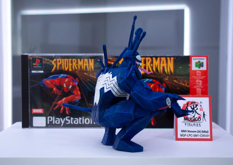 May include: A blue Venom figure with white accents and a long tongue, posed in front of a PlayStation Spider-Man game case. A red and white sign reads "MUGICO FIGURES" and "SM1 Venom (A) (Mid)".