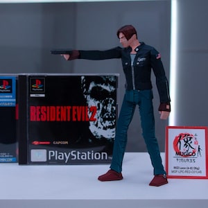 May include: A plastic action figure of Leon S. Kennedy from the Resident Evil 2 video game. The figure is holding a gun and is wearing a black jacket, blue jeans, and brown boots. The figure is standing on a white surface in front of a video game case. The case is for the PlayStation game Resident Evil 2. The case is black and has a picture of a zombie on the front. The case also has the text "Resident Evil 2" and "PlayStation" on the front. The figure is also next to a red and white card with Japanese text and the words "MUGICO FIGURES" on it.