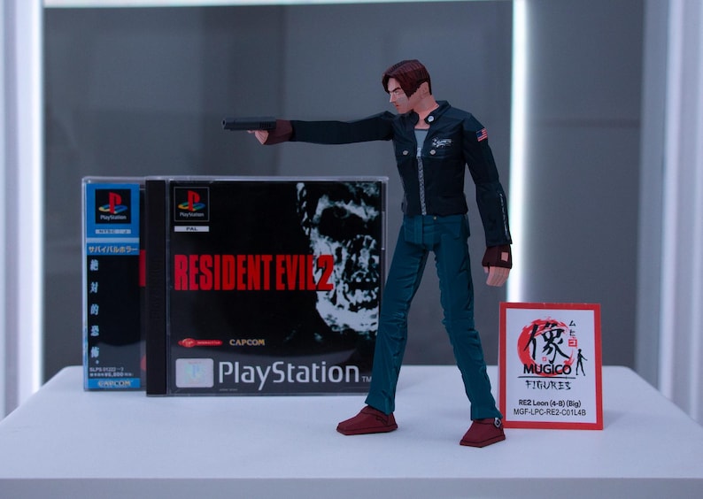 May include: A plastic action figure of Leon S. Kennedy from the Resident Evil 2 video game. The figure is holding a gun and is wearing a black jacket, blue jeans, and brown boots. The figure is standing on a white surface in front of a video game case. The case is for the PlayStation game Resident Evil 2. The case is black and has a picture of a zombie on the front. The case also has the text "Resident Evil 2" and "PlayStation" on the front. The figure is also next to a red and white card with Japanese text and the words "MUGICO FIGURES" on it.