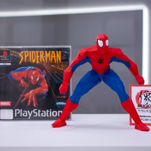 May include: A red and blue origami-style Spider-Man action figure stands on a white surface. The figure is holding a red card with Japanese text and the words "MUGICO FIGURES" and "Spiderman (A) (Big) PC-SM1-C015PB01". A PlayStation game case for Spider-Man is visible in the background.