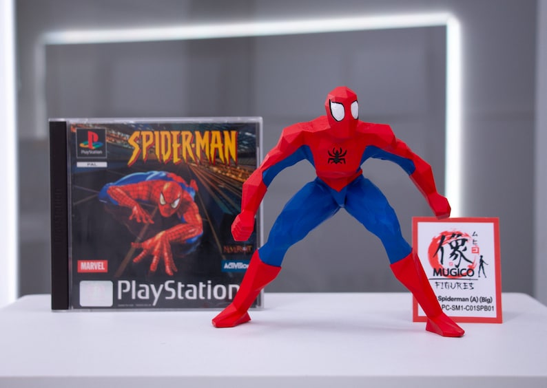 May include: A red and blue origami-style Spider-Man action figure stands on a white surface. The figure is holding a red card with Japanese text and the words "MUGICO FIGURES" and "Spiderman (A) (Big) PC-SM1-C015PB01". A PlayStation game case for Spider-Man is visible in the background.